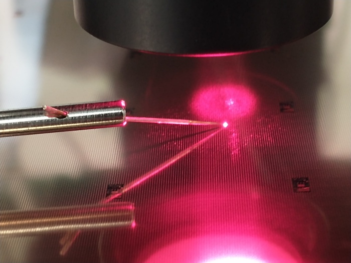 Probe needles contacting a laser die under a microscope, with the emitted spot visible on the wafer surface